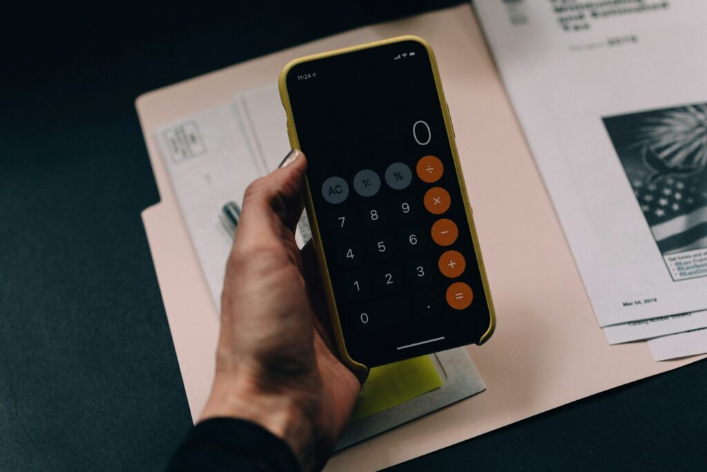 A hand holds a phone with the calculator open. Underneath the hand is an open A4 folder and documents.