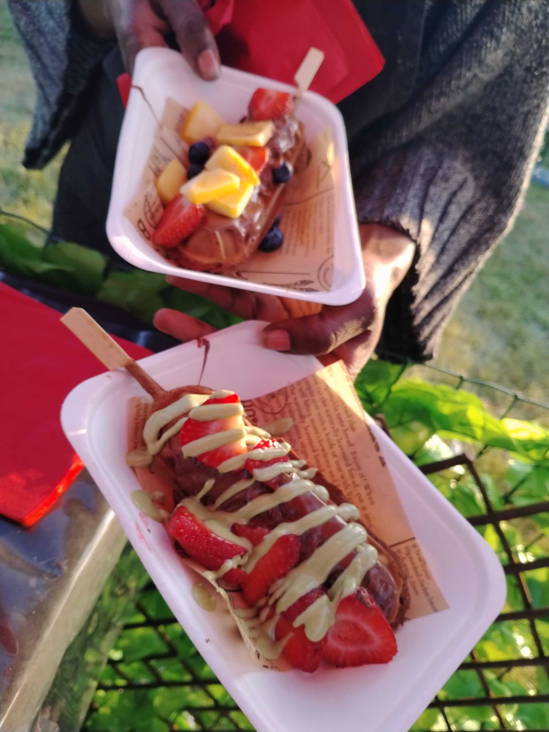 Two long waffles on popsicle sticks are on carboard trays, help by a Black woman. They're covered in sweet toppings.