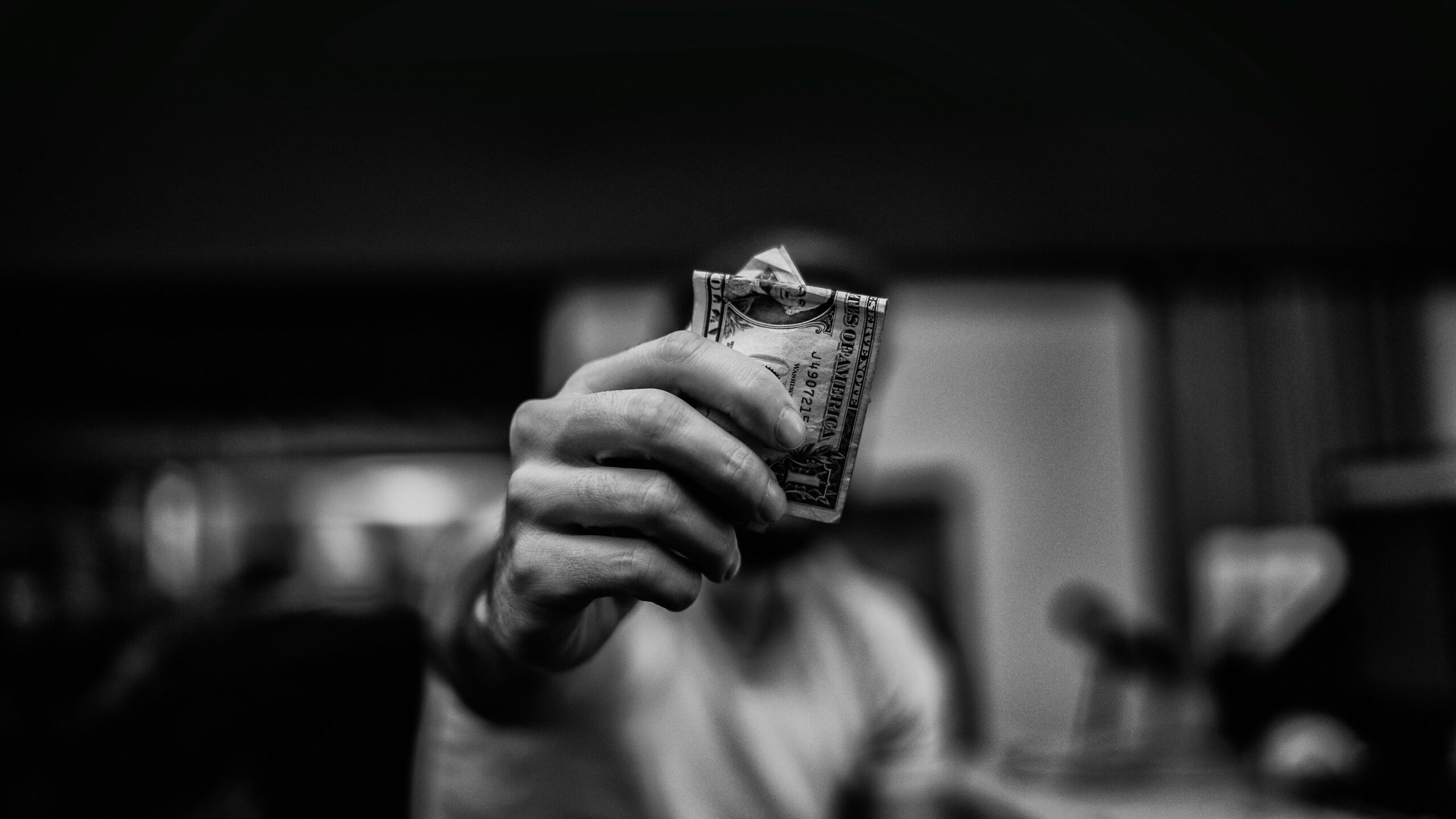 The photograph shows a close-up of a man’s hand holding a single US dollar note with a soft-focus background. The composition highlights the intricate details of the banknote against a blurred backdrop.