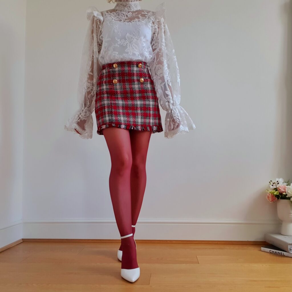 A woman stands on a wooden floor, with a white wall behind her. She's wearing a white blouse, a tartan-print mini skits, red tights, and white high heels. Her head isn't in the shot.