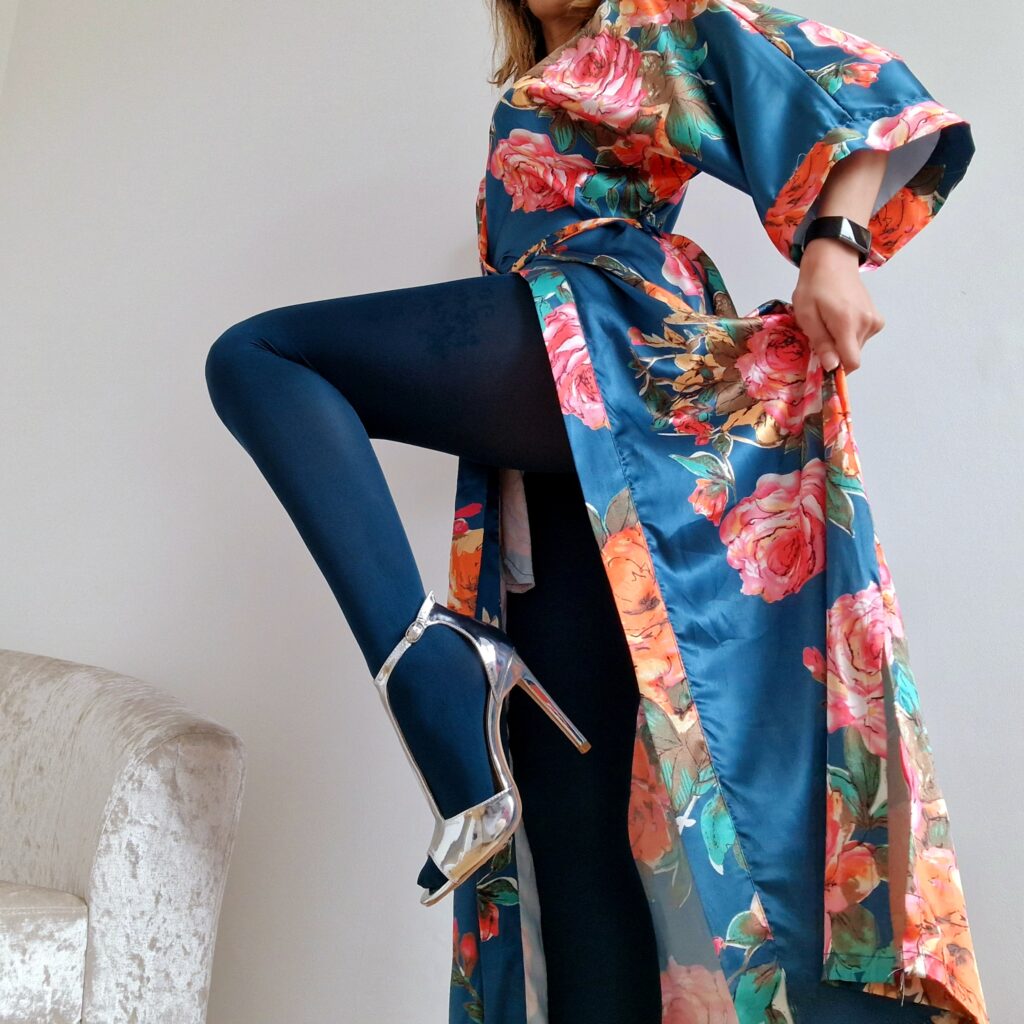 A woman stands on a wooden floor, with a white wall behind her. She's wearing dark blue tights and a colourful kimono. Her right leg is being held up, knee bent. She's wearing high heels. Her head isn't in the shot.