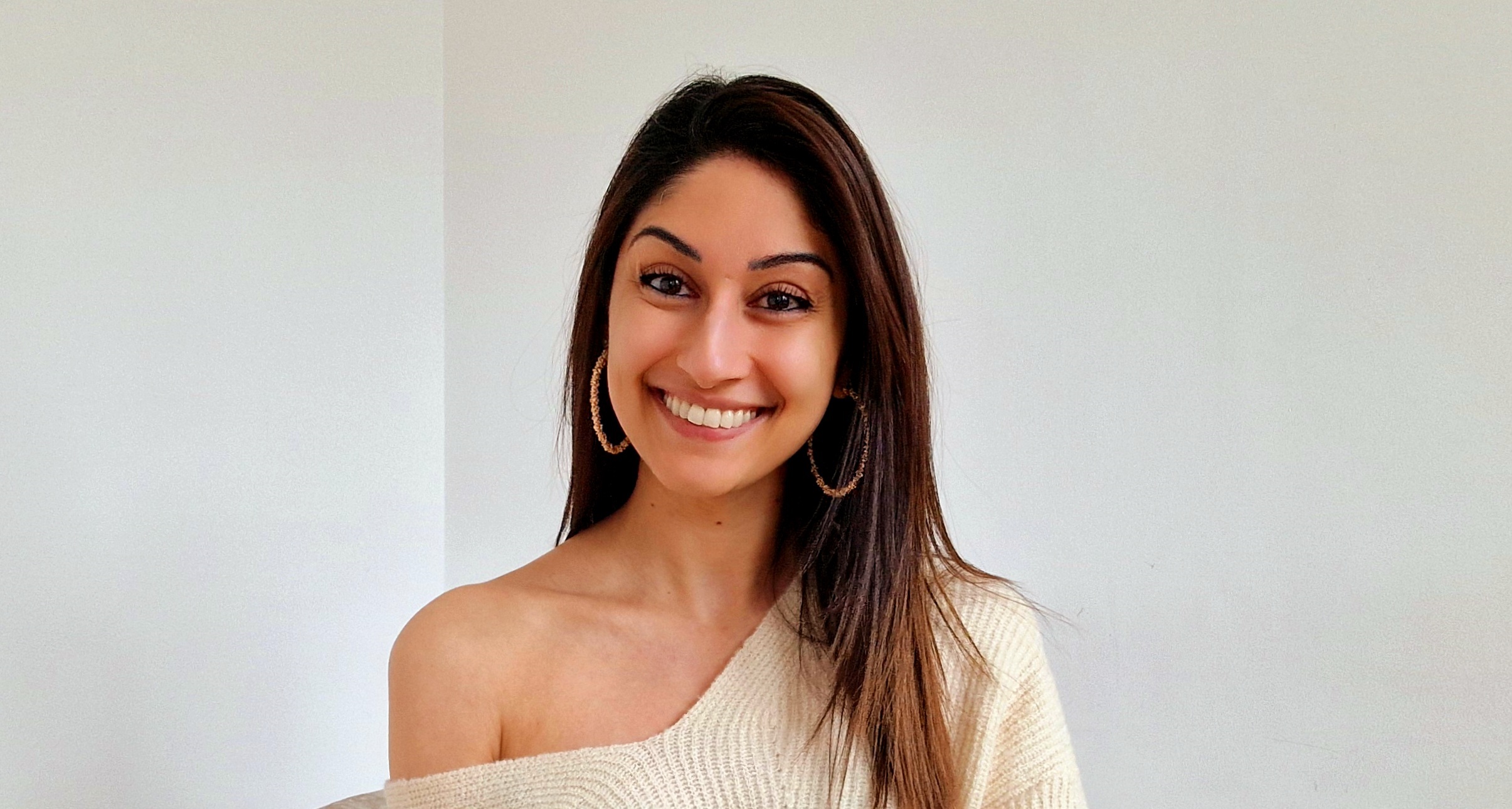 A petite slim woman with brown skin and long straight dark brown hair sits crossed legged on a grey velvet chair. She wears an off the shoulder cream sweater. She's smiling into the camera.